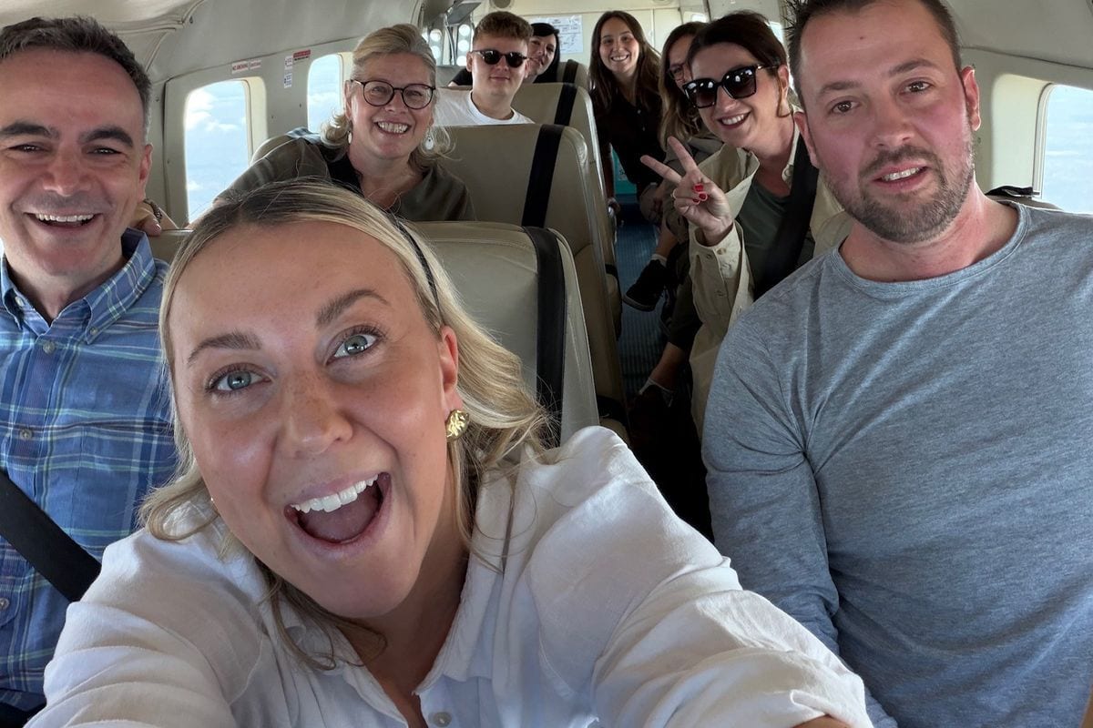 Eine Gruppe von acht Personen sitzt lächelnd in der Kabine eines kleinen Flugzeugs und schaut in die Kamera, während eine Frau im Vordergrund ein Selfie macht.