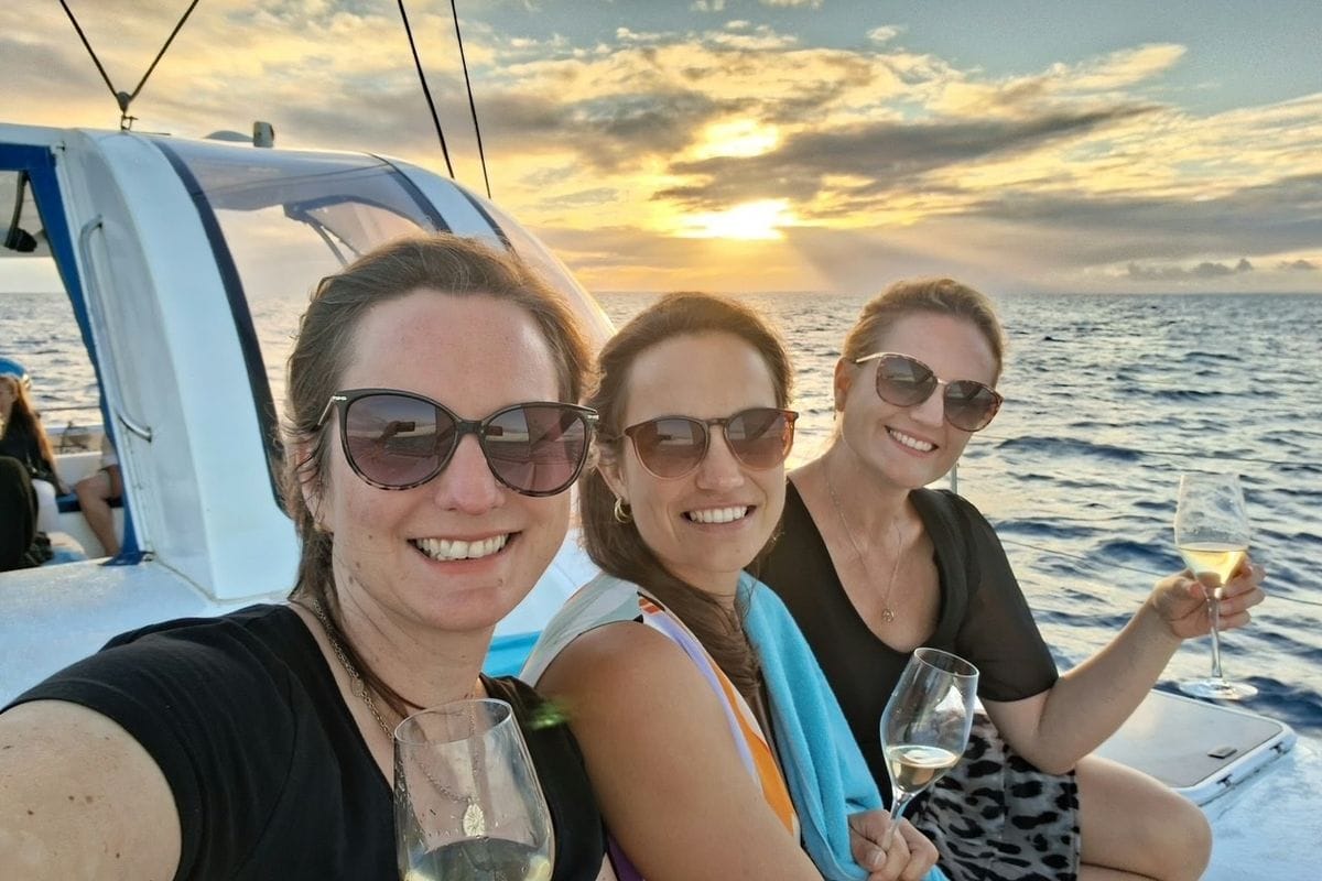 Drei lächelnde Frauen mit Sonnenbrillen sitzen auf einem Boot auf dem offenen Meer und halten Weingläser vor einem goldenen Sonnenuntergang am Horizont.