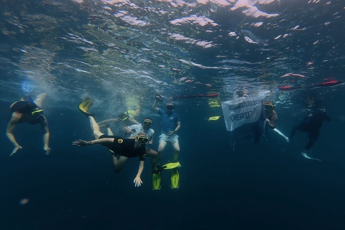 Eine Gruppe von Menschen mit Tauchmasken und gelben Flossen schwimmt unter Wasser, während eine Person eine Flagge mit der Aufschrift DERTOUR hält.