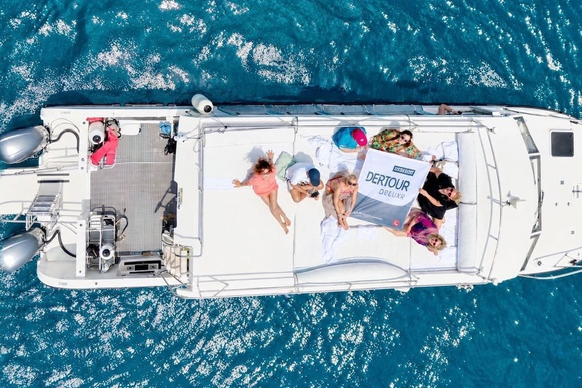 Eine Gruppe von Menschen liegt auf dem weißen Deck eines Bootes auf dem tiefblauen Meer und hält gemeinsam ein Banner mit der Aufschrift DERTOUR DELUXE.