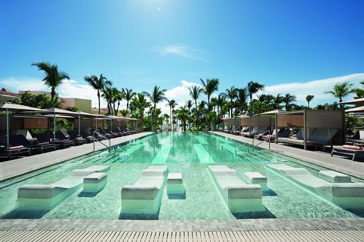 Ein langer, rechteckiger Swimmingpool mit im Wasser stehenden Liegen, umgeben von Palmen, Sonnenschirmen und Liegeflächen unter einem klaren, blauen Himmel.