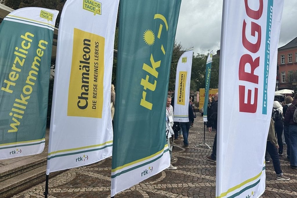 Mehrere aufgestellte Werbefahnen mit Logos von rtk, Chamäleon und ERGO Reiseversicherung stehen auf einem gepflasterten Platz vor einem historischen Gebäude.