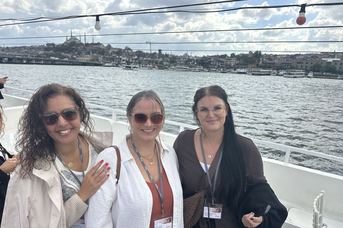 Drei lächelnde Frauen stehen auf einem Boot vor der Kulisse einer Stadt am Wasser, tragen Namensschilder an Umhängebändern und blicken in die Kamera.