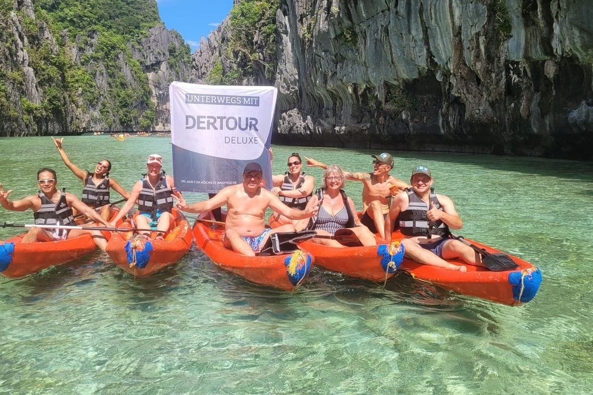 Eine Gruppe von Menschen in orangefarbenen Kajaks auf türkisfarbenem Wasser vor steilen Felswänden, eine Person hält eine Dertour Deluxe Flagge in die Höhe.