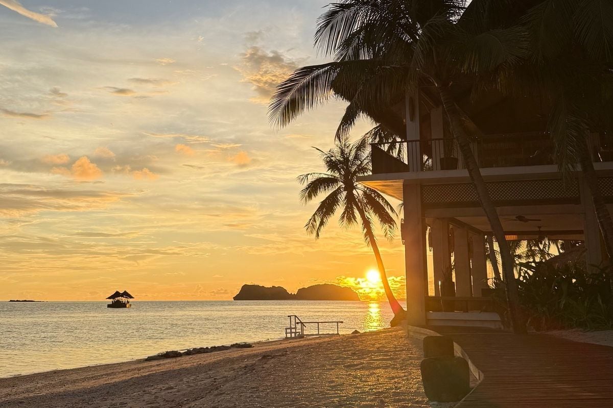 Ein goldener Sonnenuntergang über dem Meer mit Palmen, einem hölzernen Steg und einem Gebäude am Strand vor einer Inselkulisse in einer tropischen Region.