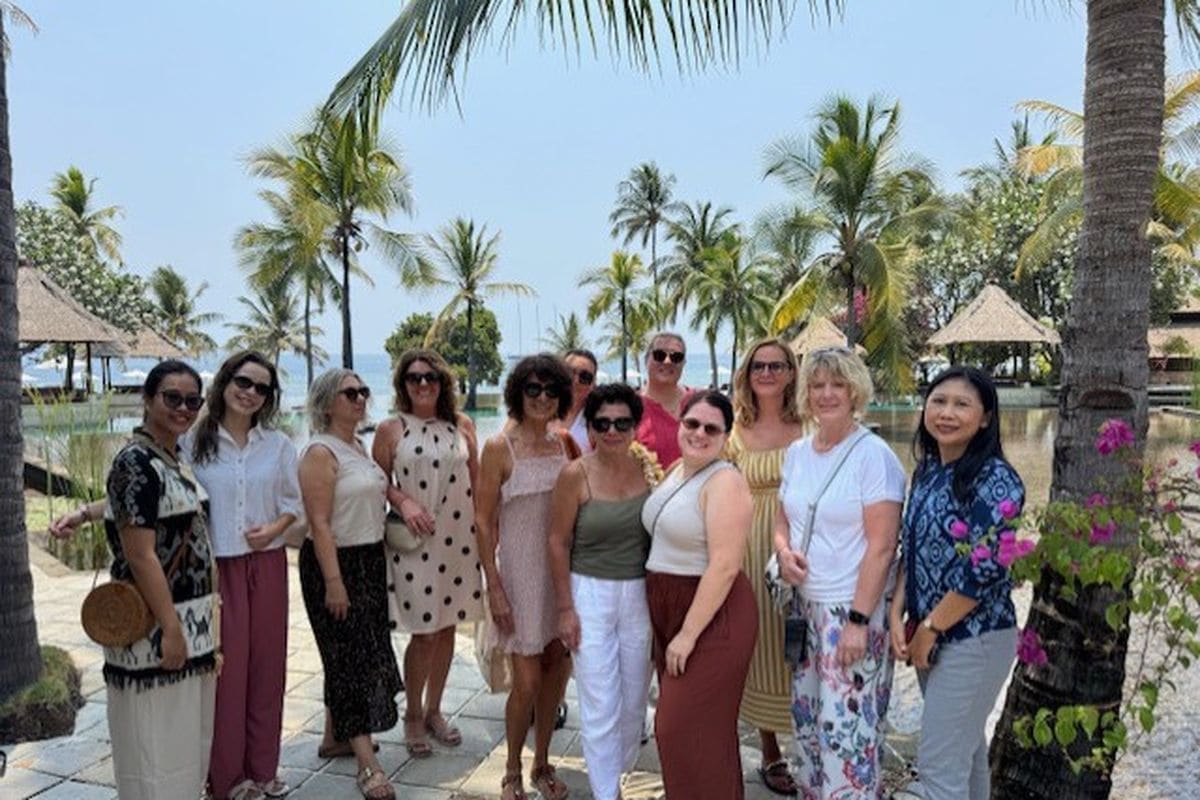 Eine Gruppe von elf Frauen steht lächelnd in einer tropischen Hotelanlage mit Palmen und Wasserbecken im Hintergrund für ein gemeinsames Foto zusammen.