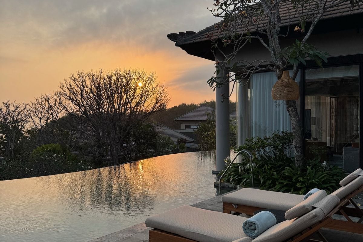 Ein Infinity-Pool vor einer Villa bei Sonnenuntergang mit zwei Liegen im Vordergrund, umgeben von Bäumen und einer hügeligen Landschaft im Hintergrund.
