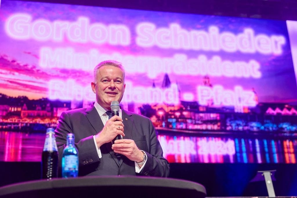 Ein Mann im dunklen Anzug spricht in ein Mikrofon vor einer großen Leinwand, auf der der Name Gordon Schnieder und Rheinland-Pfalz in Neonoptik stehen.