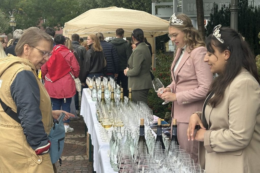 Zwei Frauen mit Diademen stehen hinter einem langen Tisch mit Weingläsern und Flaschen, während eine Besucherin im Vordergrund das Angebot betrachtet.