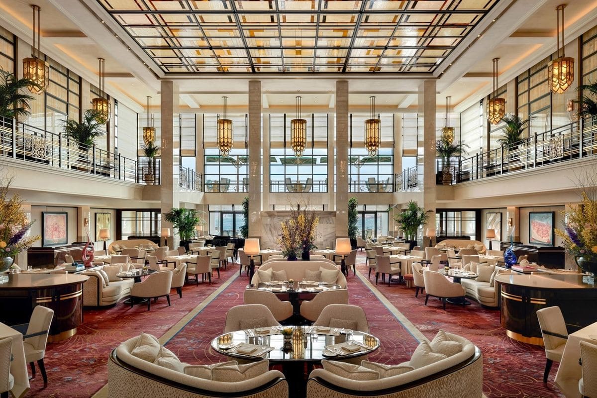 Ein weitläufiger, eleganter Speisesaal mit hohen Decken, Art-déco-Kronleuchtern, cremefarbenen Polstermöbeln und einem gemusterten roten Teppichboden.