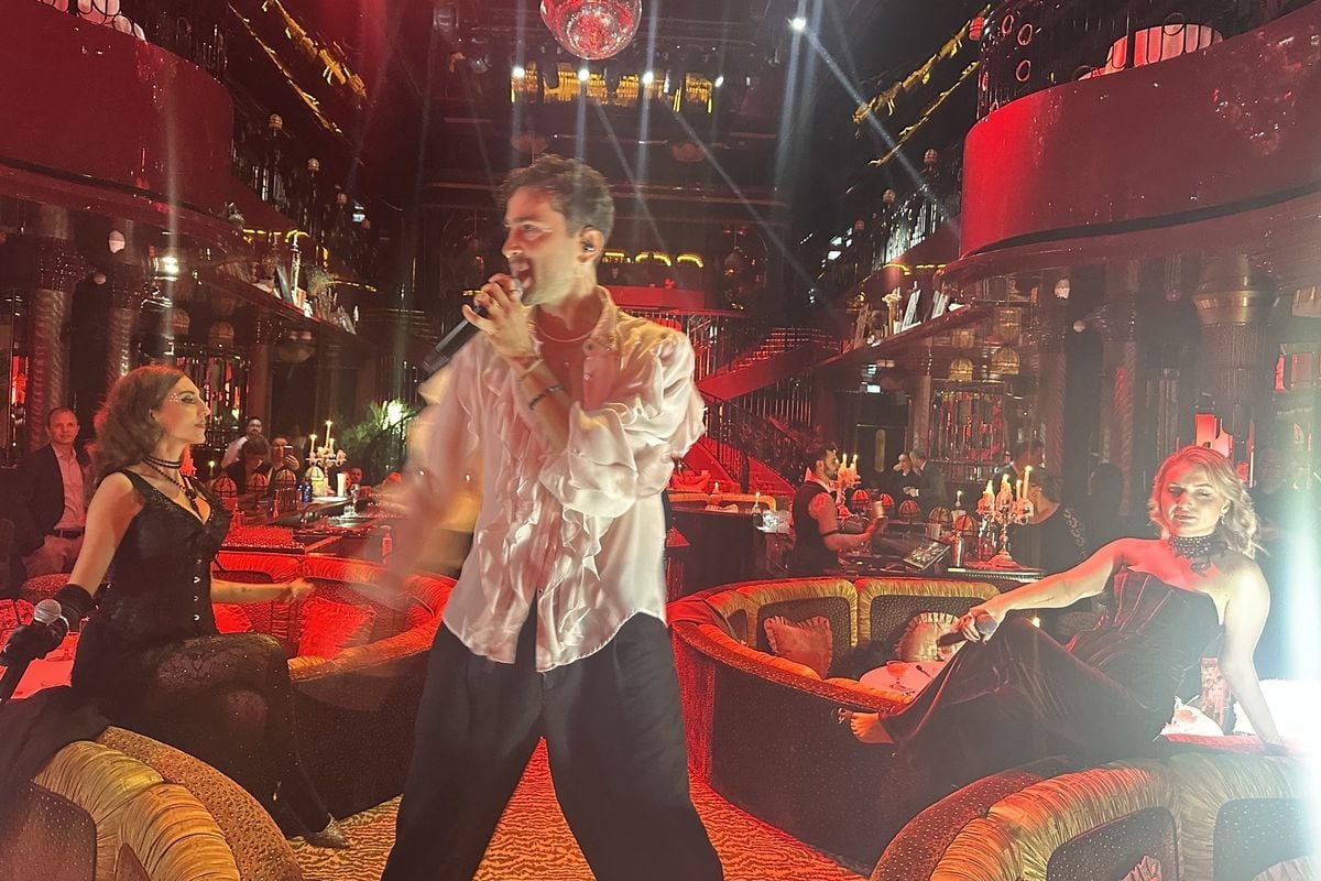 Ein Mann mit weißem Rüschenhemd singt in einem prunkvoll gestalteten, rot beleuchteten Club, flankiert von zwei Frauen in schwarzen Outfits auf Sofas.