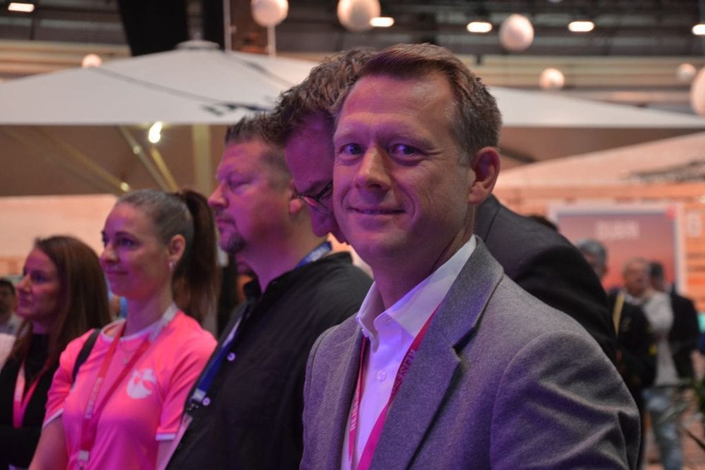 Ein Mann im grauen Sakko und weißem Hemd blickt in die Kamera, während neben ihm weitere Personen in einer Halle mit großen Sonnenschirmen stehen.