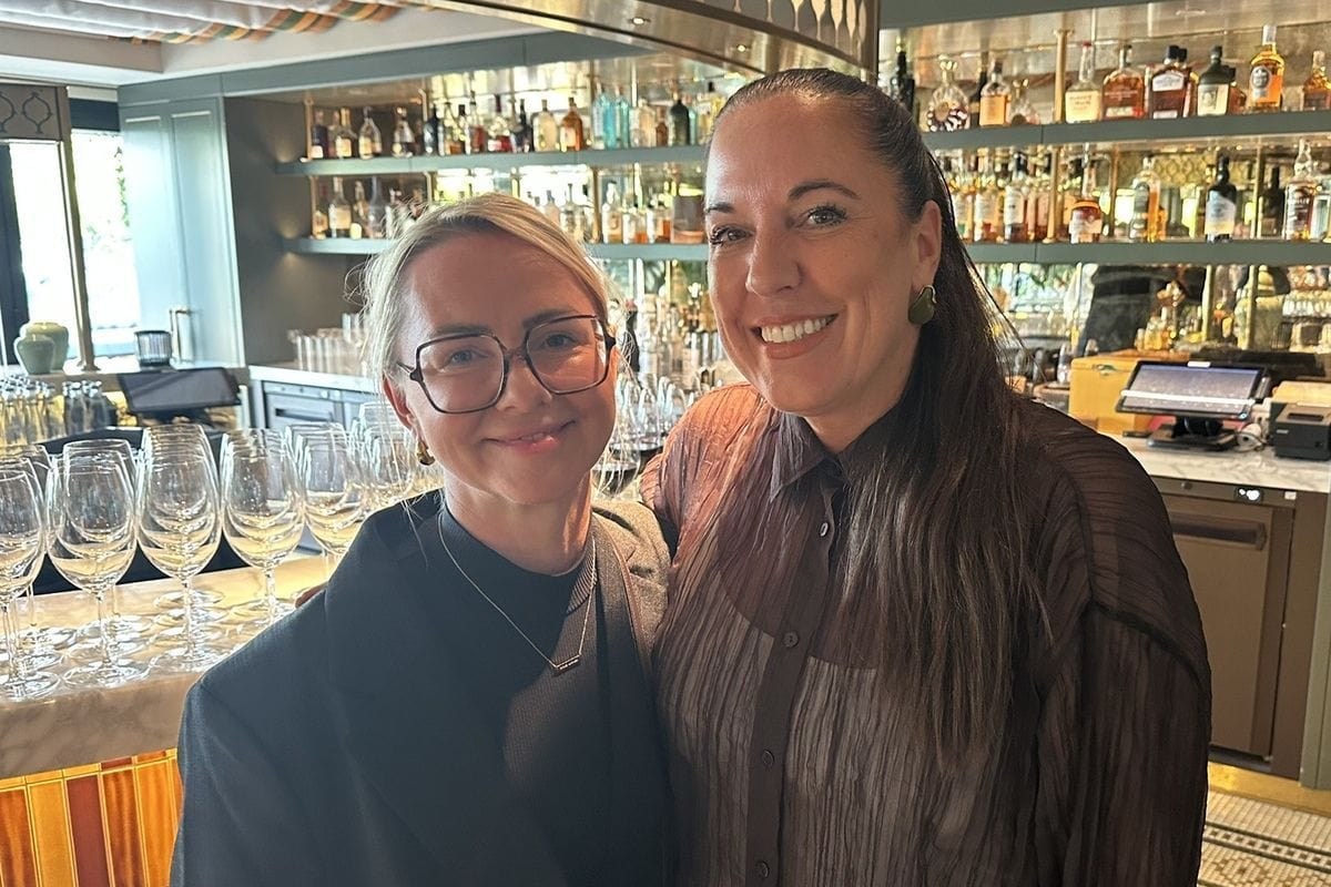 Zwei lächelnde Frauen stehen eng beieinander vor einer Bar mit beleuchteten Regalen voller Flaschen und leeren Weingläsern auf einem Tresen im Vordergrund.