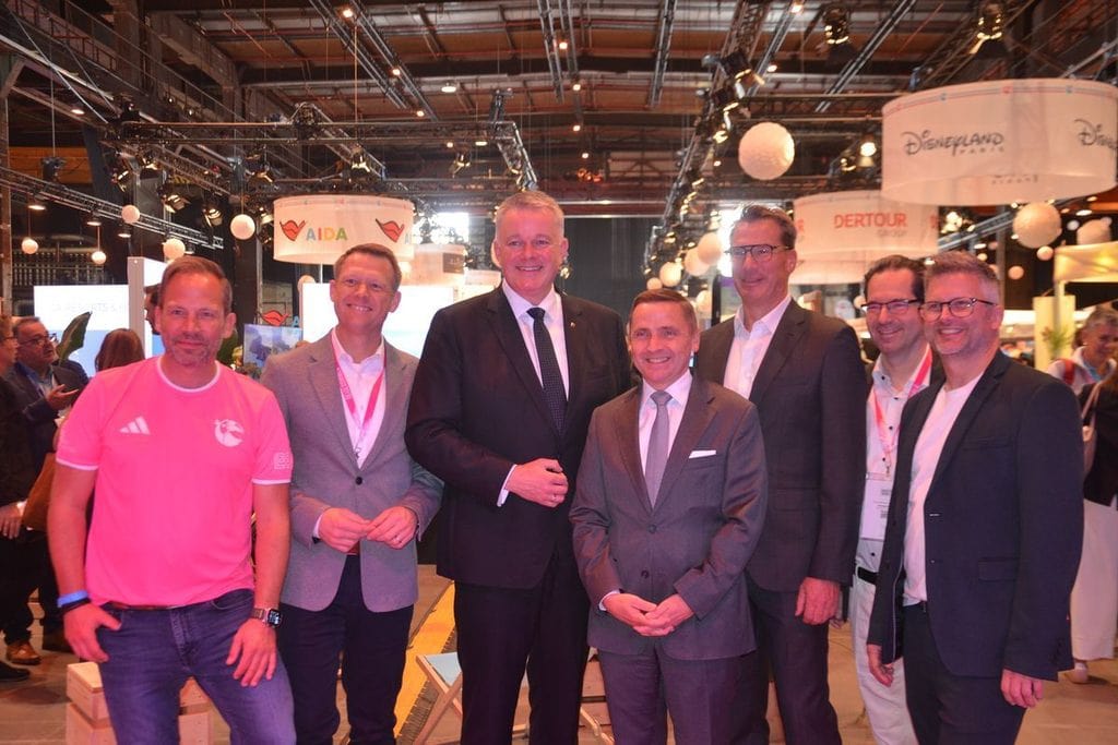 Eine Gruppe von sechs Männern steht in einer Messehalle vor verschiedenen Firmenlogos wie AIDA und Dertour und lächelt in die Kamera für ein Gruppenfoto.