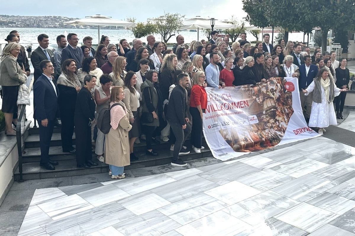 Eine große Gruppe von Menschen steht auf einer Steintreppe vor dem Wasser und hält gemeinsam ein großes Banner mit der Aufschrift Herzlich Willkommen.