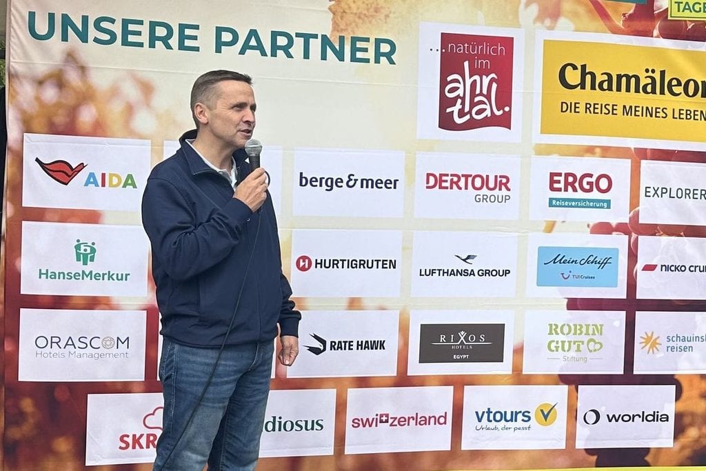 Ein Mann in einer dunkelblauen Jacke und Jeans spricht vor einer großformatigen Sponsorenwand mit verschiedenen Firmenlogos in ein Mikrofon.