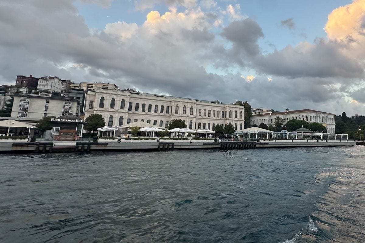 Blick vom Wasser auf das Four Seasons Hotel am Bosporus in Istanbul mit einer langen Uferpromenade, weißen Sonnenschirmen und bewölktem Abendhimmel.