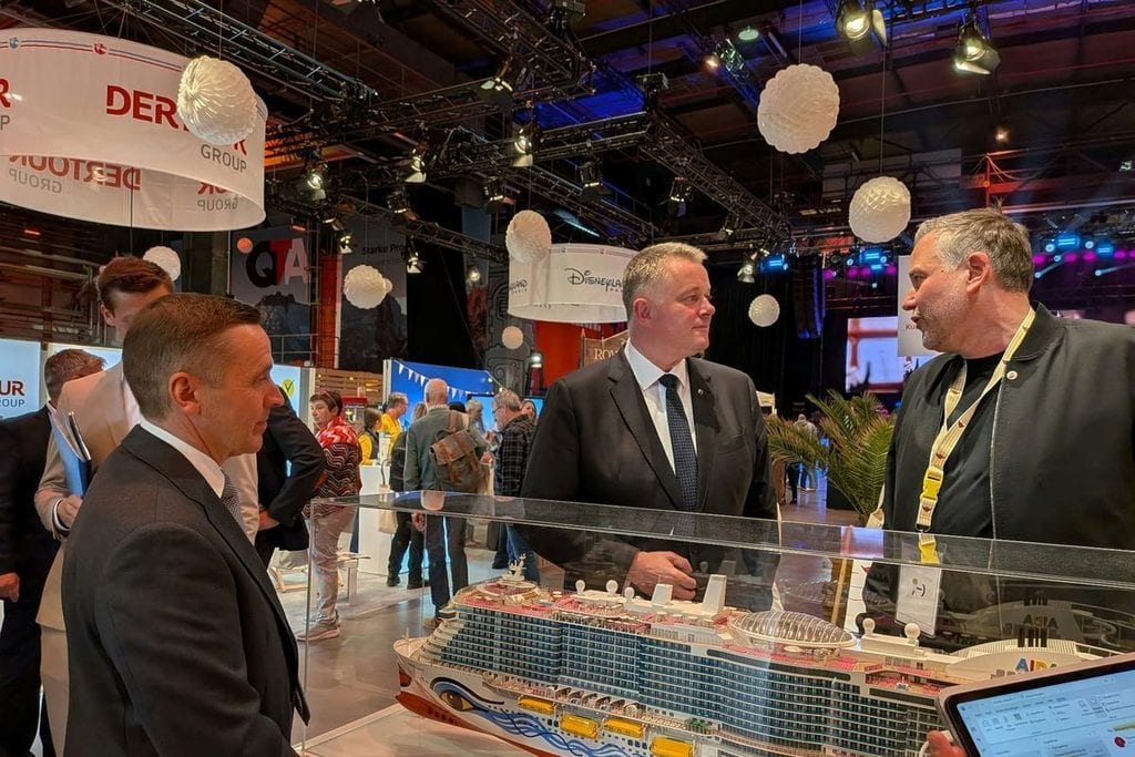 Drei Männer in formeller Kleidung stehen auf einer Messe vor einem Modell eines AIDA-Kreuzfahrtschiffes und führen ein Gespräch in einer Messehalle.