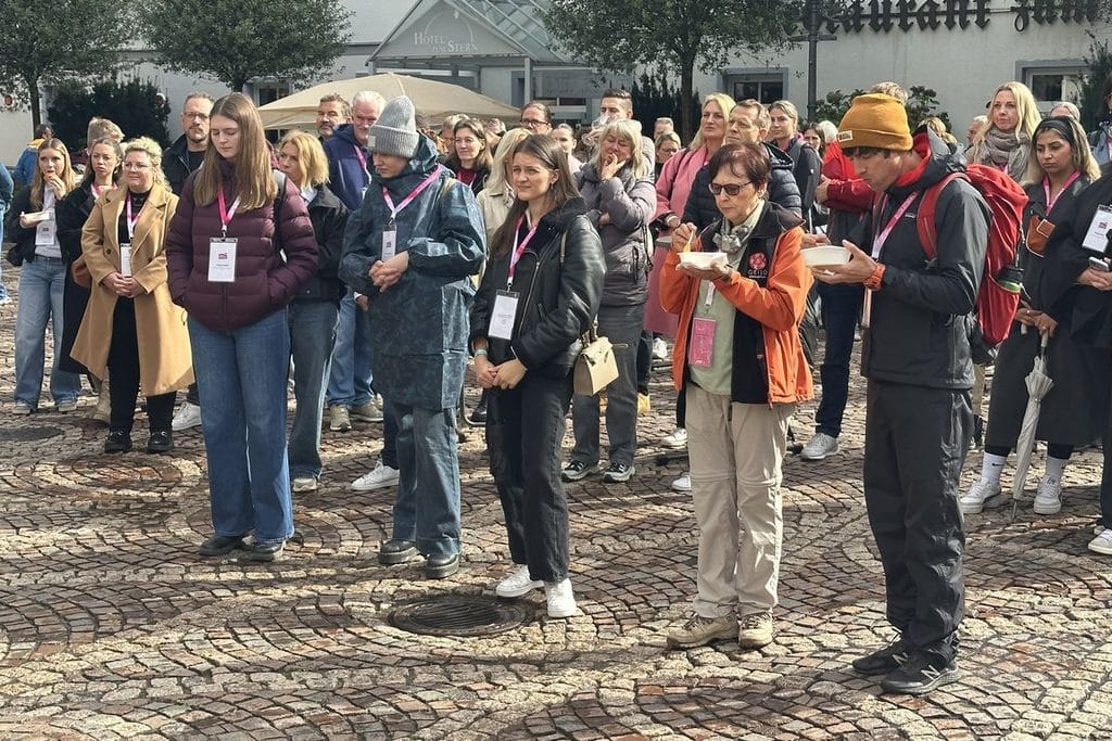 Eine Gruppe von Menschen steht auf einem gepflasterten Platz vor einem Hotelgebäude, einige Personen essen aus kleinen Schalen, viele tragen Namensschilder.