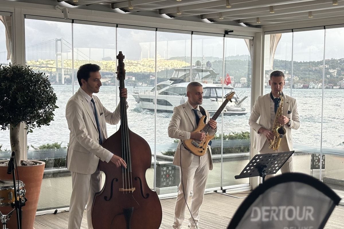 Drei Musiker in hellen Anzügen spielen Kontrabass, E-Gitarre und Saxophon vor einer Glasfront mit Blick auf eine Yacht und eine Hängebrücke am Wasser.