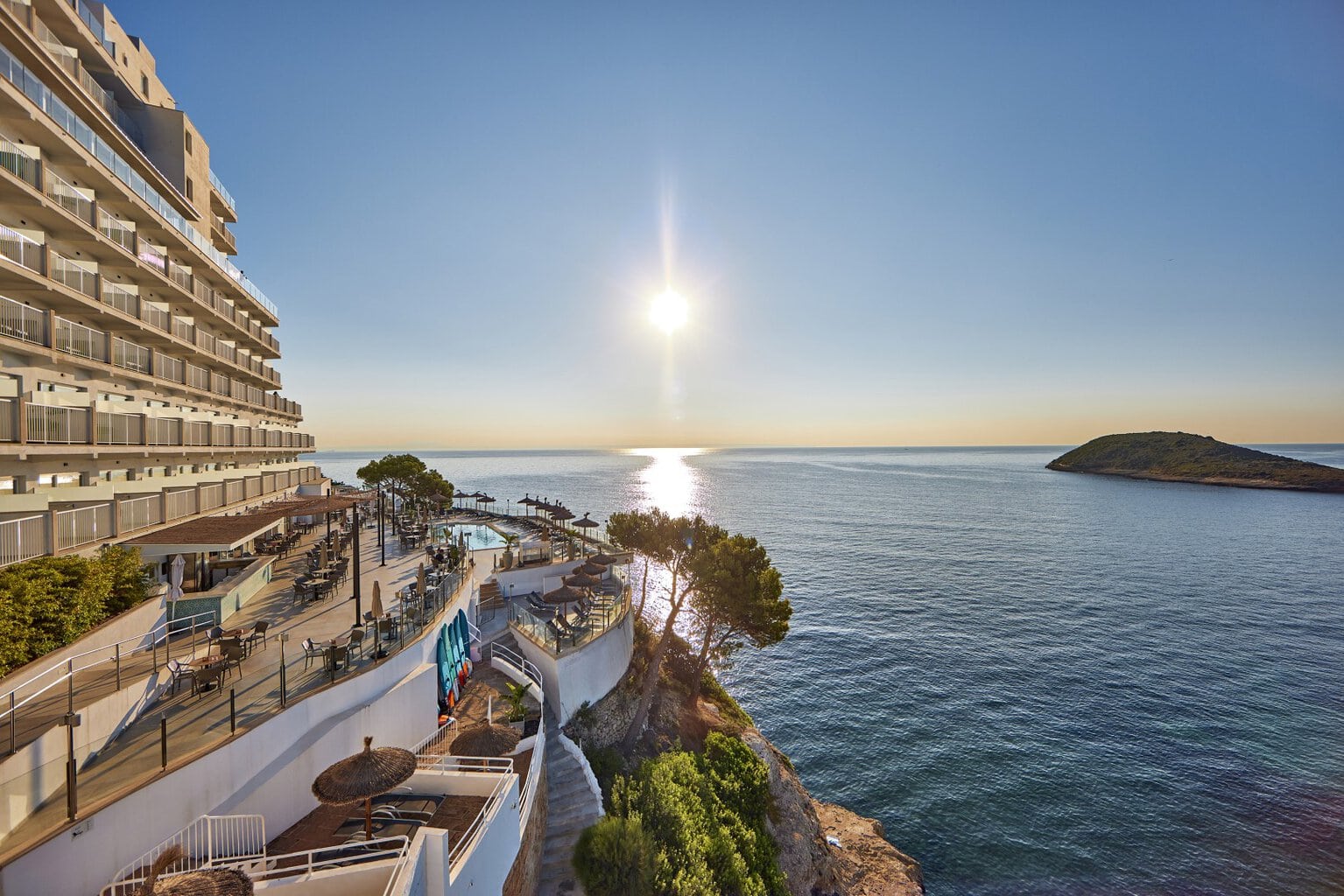 Blick auf ein Hotelgebäude am Meer mit einem Poolbereich und einer kleinen Insel im Hintergrund, bei Sonnenuntergang.