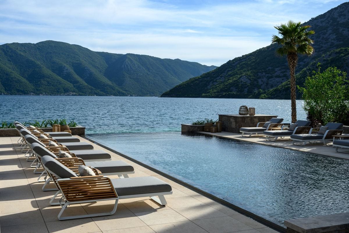 Eine Reihe von Sonnenliegen auf einer Terrasse an einem Infinity-Pool mit Blick auf eine weite Bucht, umgeben von hohen, grünen Bergen unter blauem Himmel.