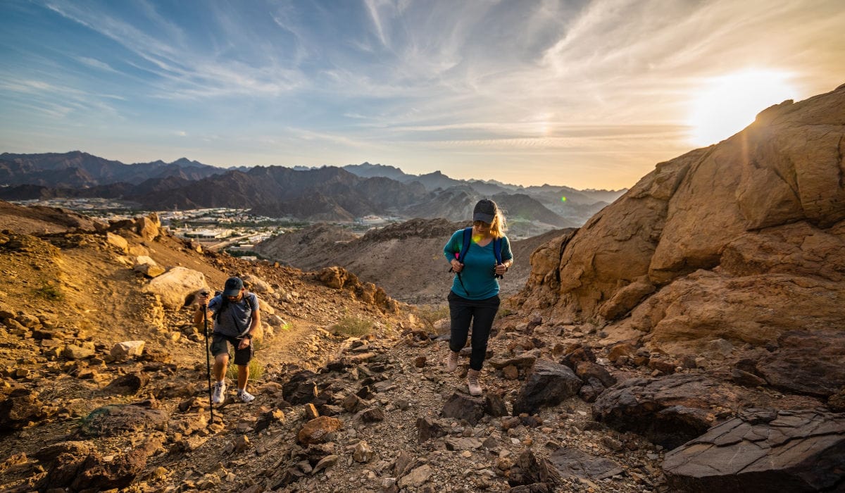 Zwei Wanderer mit Rucksäcken steigen einen steinigen Pfad in einer felsigen Berglandschaft bei Sonnenuntergang hinab, im Hintergrund liegt ein Tal.