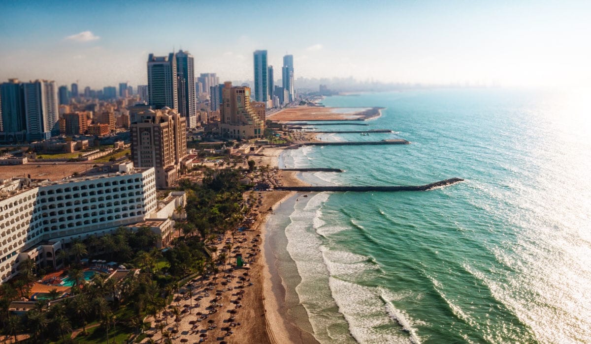 Luftaufnahme einer Küstenlinie mit einer modernen Skyline, einem Sandstrand mit Sonnenschirmen und mehreren ins Meer ragenden Wellenbrechern bei Tageslicht.