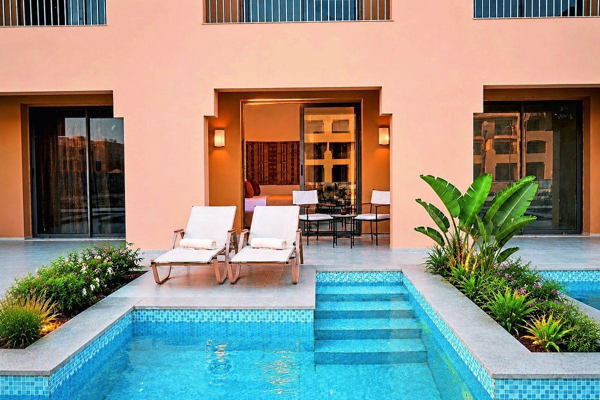 Zwei weiße Sonnenliegen stehen auf einer Terrasse vor einem Hotelzimmer mit Poolzugang, flankiert von grünen Pflanzen und einem blau gefliesten Pool.