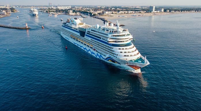Ein großes AIDA-Kreuzfahrtschiff fährt bei ruhigem Wasser aus einer Hafeneinfahrt hinaus, im Hintergrund ist eine Küstenlinie mit Bebauung zu erkennen.