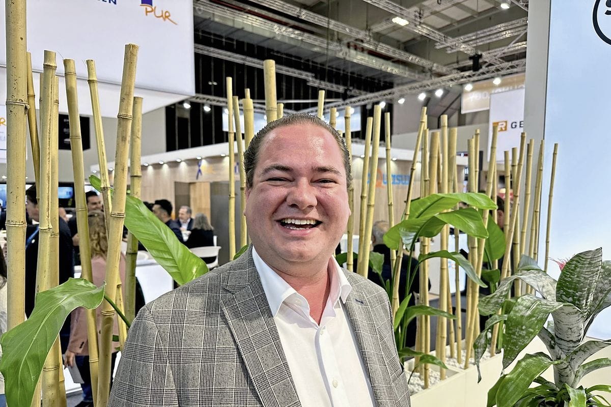 Ein lächelnder Mann in einem karierten Sakko und weißem Hemd steht vor einer dekorativen Kulisse aus vertikalen Bambusstangen und grünen Zimmerpflanzen.