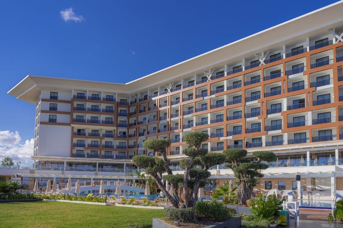 Ein modernes, mehrstöckiges Hotelgebäude mit markanter braun-weißer Fassade, Balkonen und einer weitläufigen Außenanlage mit Pool und gepflegtem Garten.