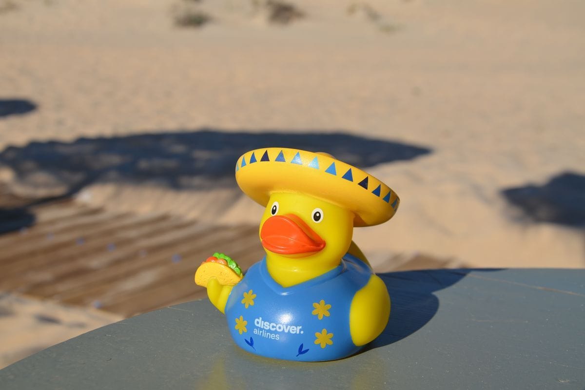 Eine gelbe Quietscheente mit Sombrero und blauem Hemd mit Discover Airlines Logo hält einen Taco in der Hand, platziert auf einem Tisch vor Sandstrand.