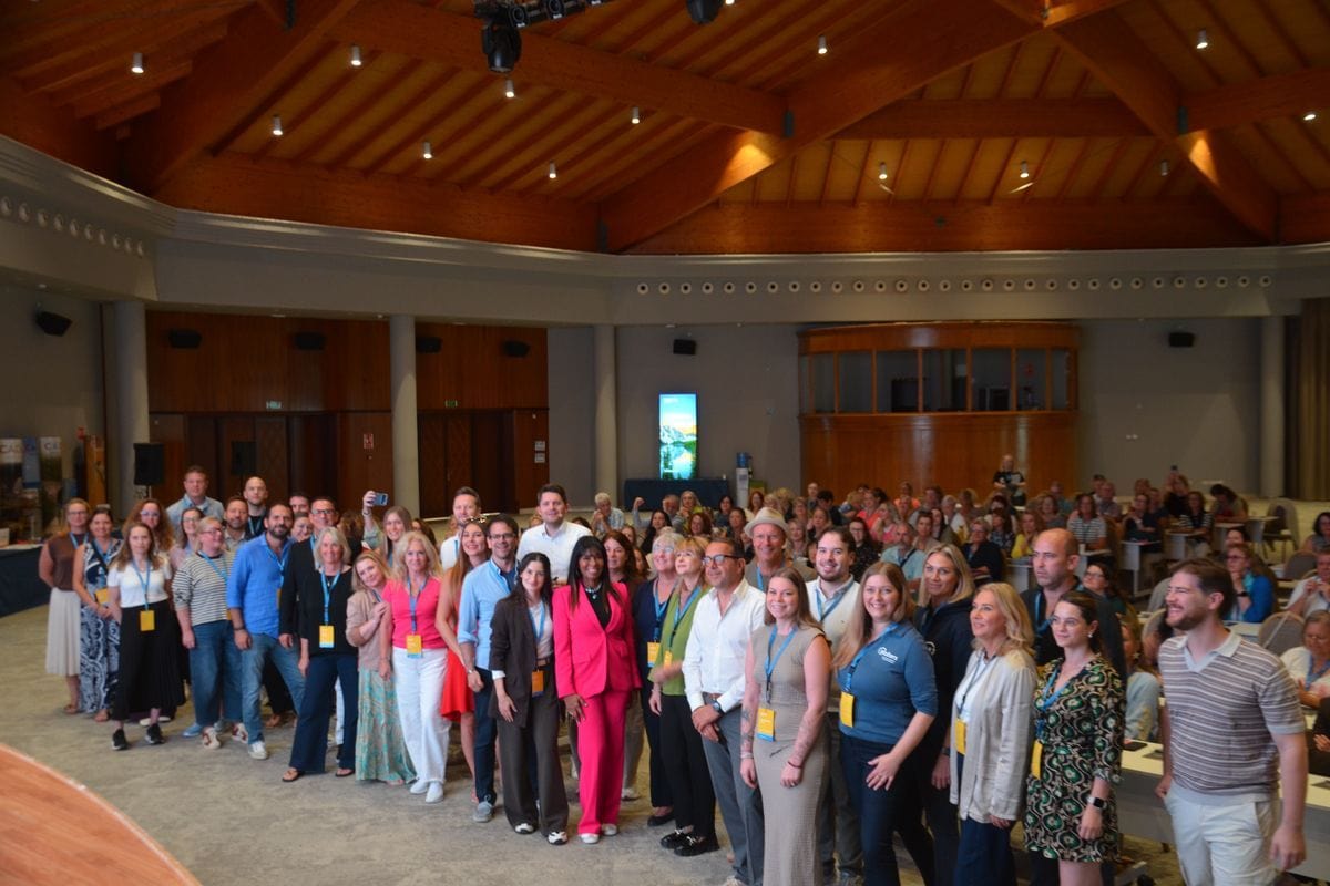 Eine große Gruppe von Menschen steht in einem weitläufigen Konferenzsaal mit hoher Holzdecke für ein Gruppenfoto zusammen und blickt in die Kamera.