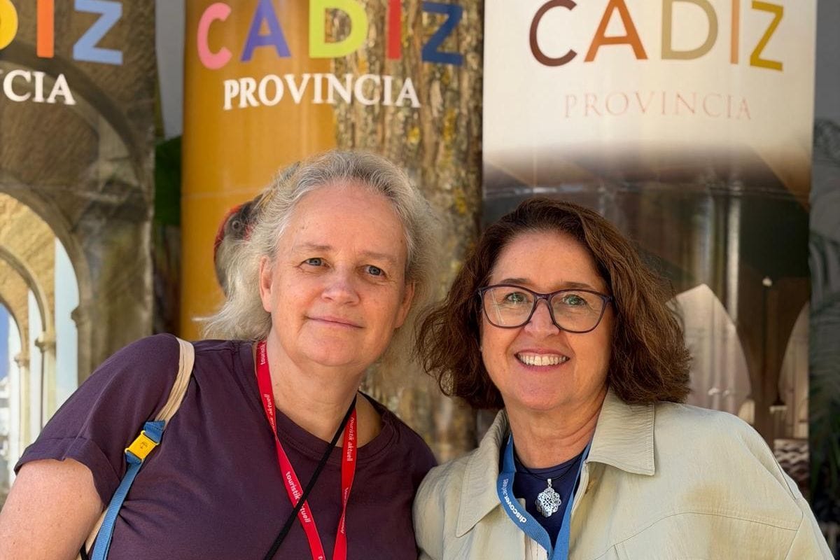 Zwei Frauen stehen nebeneinander vor Werbebannern mit der Aufschrift Cadiz Provincia. Beide tragen Schlüsselbänder um den Hals und lächeln in die Kamera.