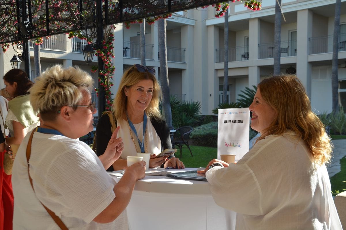 Drei Frauen stehen im Freien an einem Stehtisch mit einem Schild der Firma Viajes Karisma und unterhalten sich in einer Hotelanlage mit Palmen im Hintergrund.