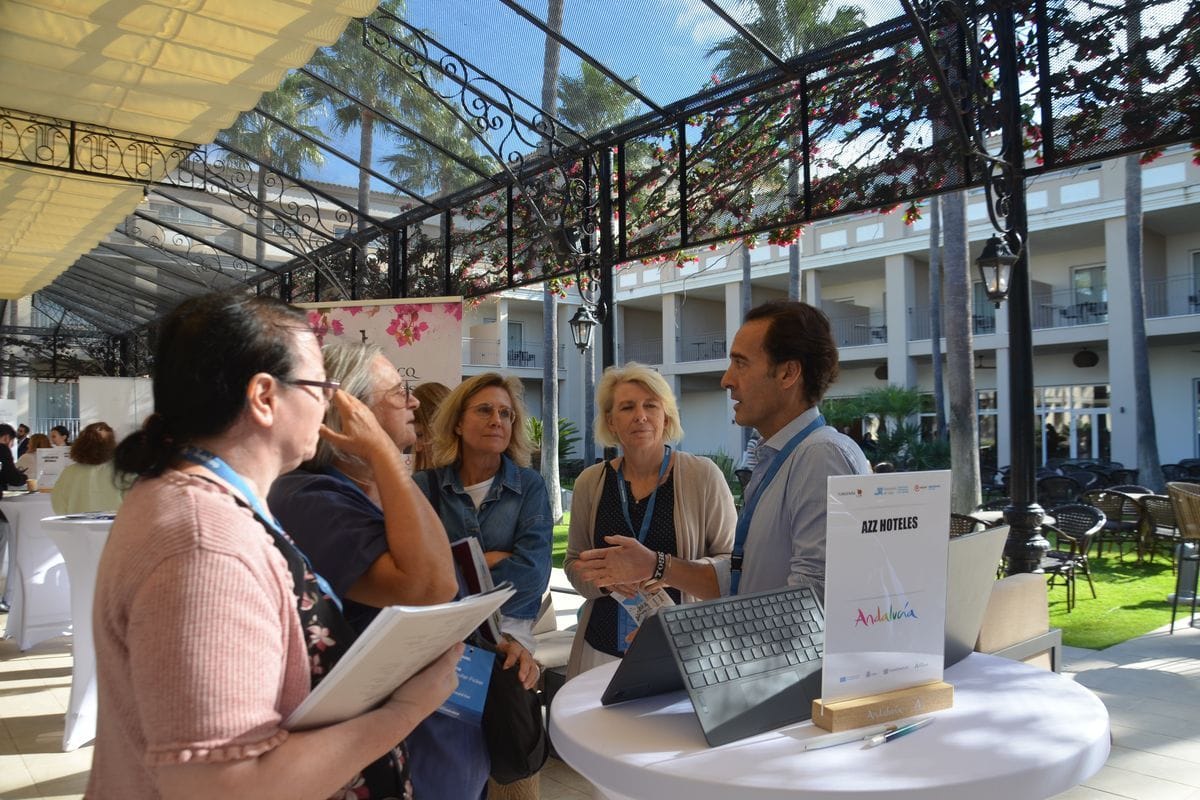 Eine Gruppe von vier Personen steht an einem runden Stehtisch mit Laptop und einem Aufsteller von AZZ HOTELES in einem überdachten Außenbereich eines Hotels.