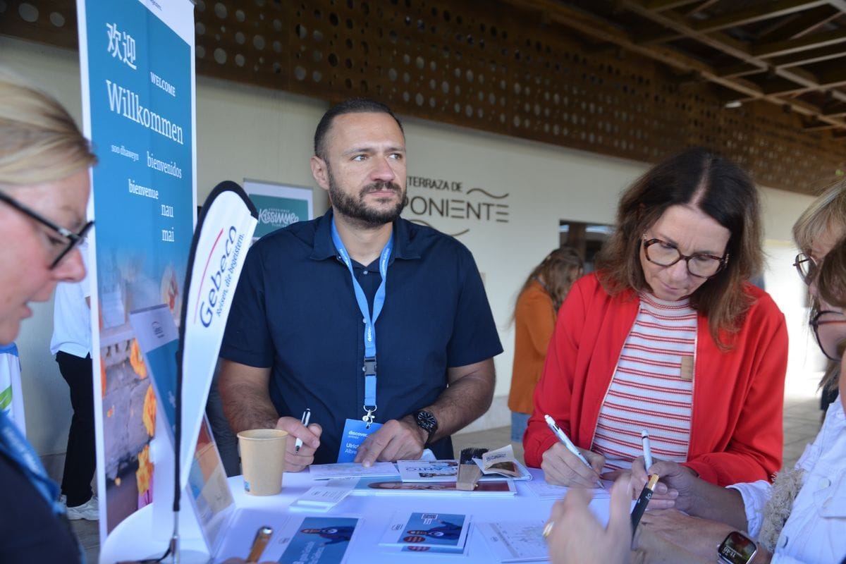 Ein Mann mit Bart und blauem Hemd steht an einem Messestand, während zwei Frauen mit Brillen Dokumente auf einem runden Tisch ausfüllen und unterschreiben.