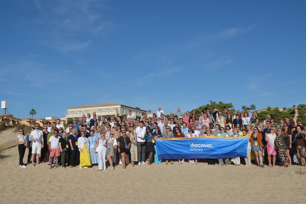 Eine große Gruppe von Menschen steht an einem sonnigen Strand vor einem Hotel und hält gemeinsam ein blaues Banner mit der Aufschrift discover airlines.