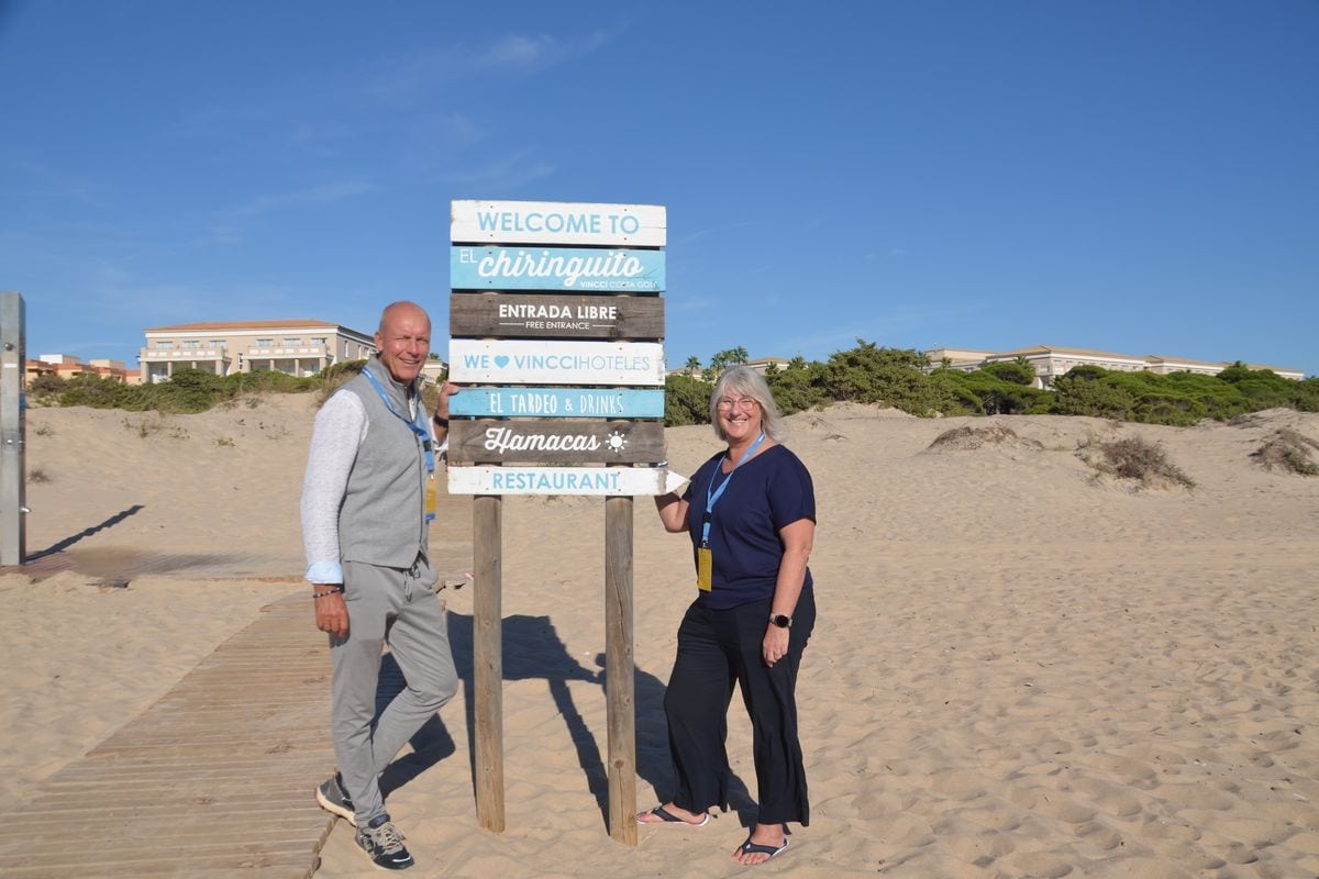Ein Mann und eine Frau stehen an einem sonnigen Tag am Sandstrand neben einem hölzernen Wegweiser mit der Aufschrift Welcome to El Chiringuito Vincci Hoteles.