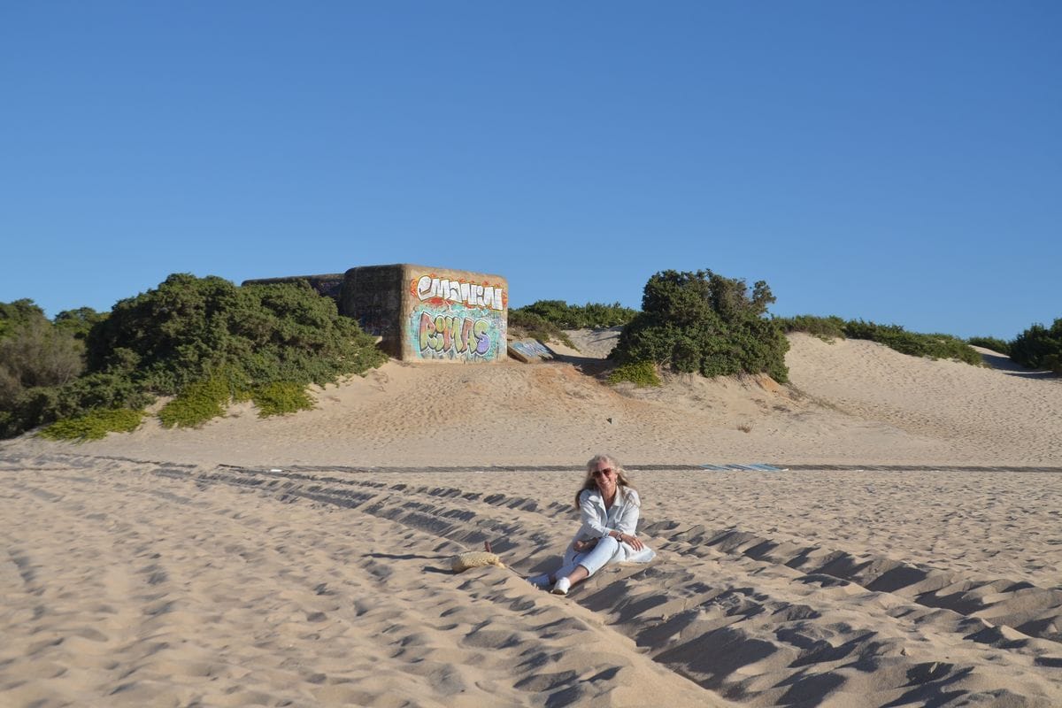 Eine Frau mit langem hellem Haar sitzt in heller Kleidung auf einem sandigen Weg vor einem mit Graffiti verzierten Betonbunker in einer Dünenlandschaft.