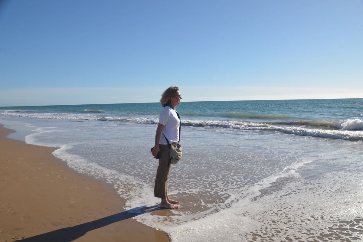 Eine Frau mit blondem Haar, weißem T-Shirt und beiger Hose steht barfuß im flachen Wasser an einem sonnigen Sandstrand und blickt auf das offene Meer hinaus.