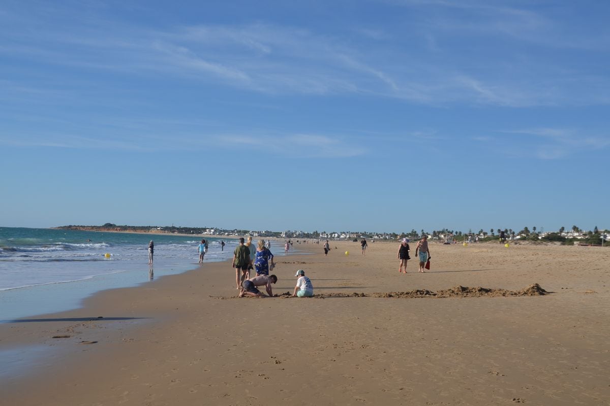 Ein weiter Sandstrand an einem sonnigen Tag mit blauem Himmel, an dem mehrere Personen spazieren gehen oder im Sand spielen, während das Meer sanft brandet.