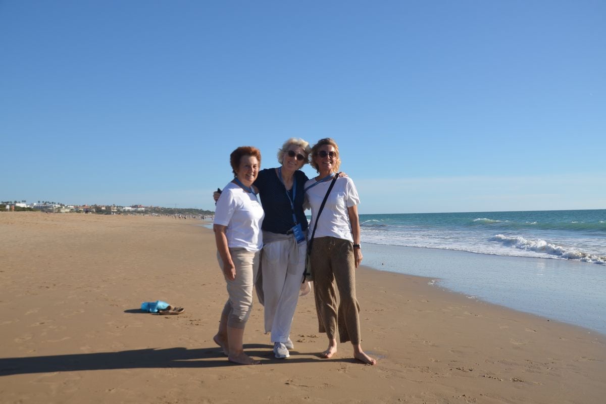 Drei Frauen stehen Arm in Arm an einem sonnigen Sandstrand am Meer, tragen Freizeitkleidung und Sonnenbrillen und blicken gemeinsam in die Kamera.