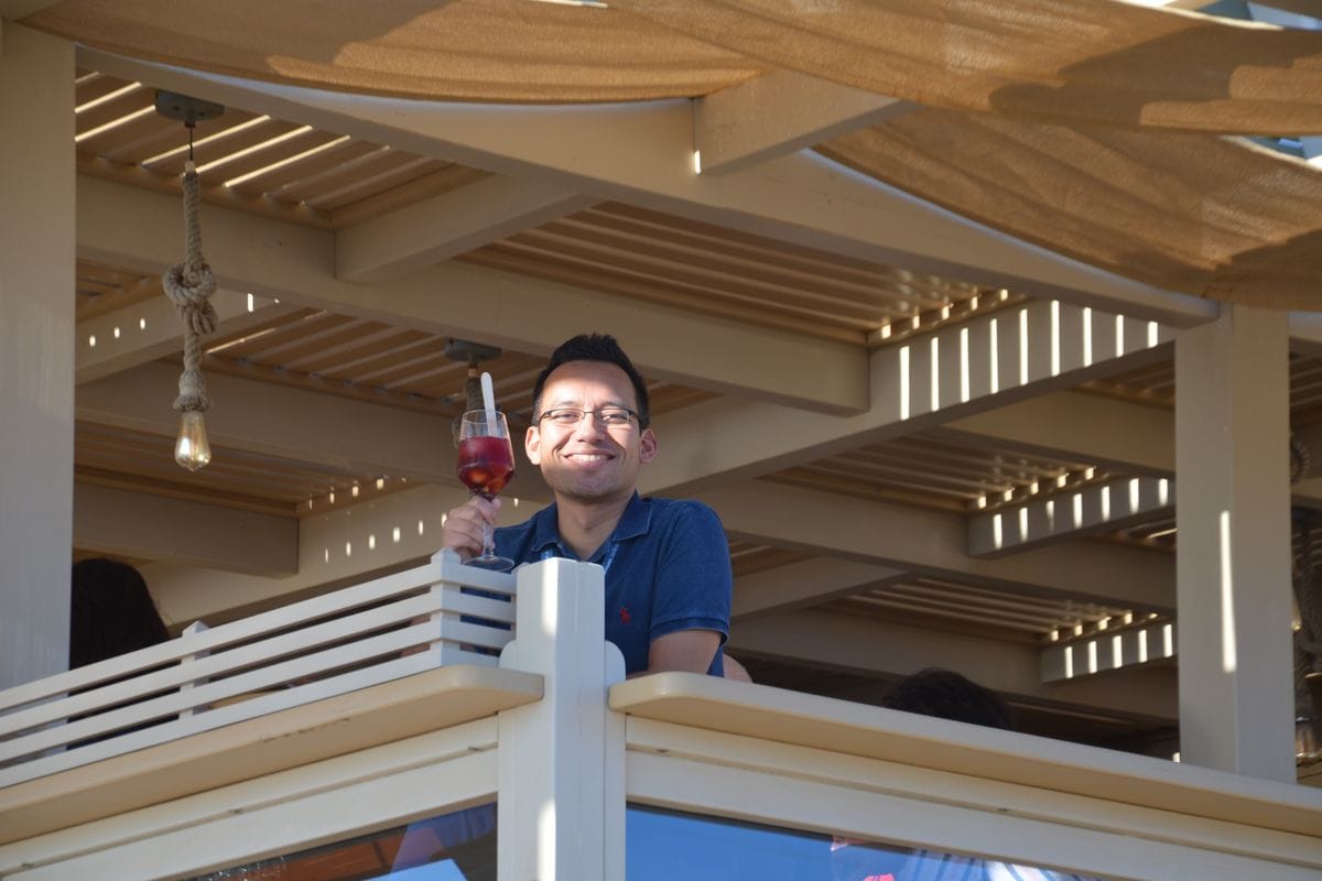 Ein lächelnder Mann mit Brille und blauem Poloshirt sitzt auf einer schattigen Terrasse und hält ein Glas mit einem rötlichen Getränk in der Hand.