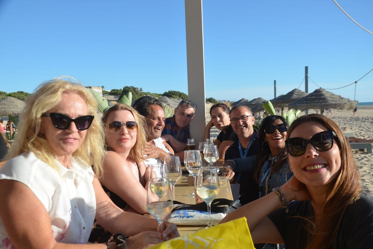 Eine Gruppe von sieben Personen sitzt an einem langen Tisch in einem Strandrestaurant unter einem Sonnendach und hält Weingläser in den Händen.