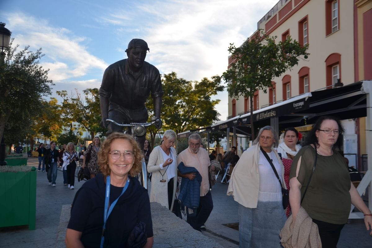 Eine Gruppe von Menschen geht auf einem sonnigen Platz an einer lebensgroßen Bronzestatue eines Radfahrers vorbei, im Hintergrund sind Gebäude zu sehen.