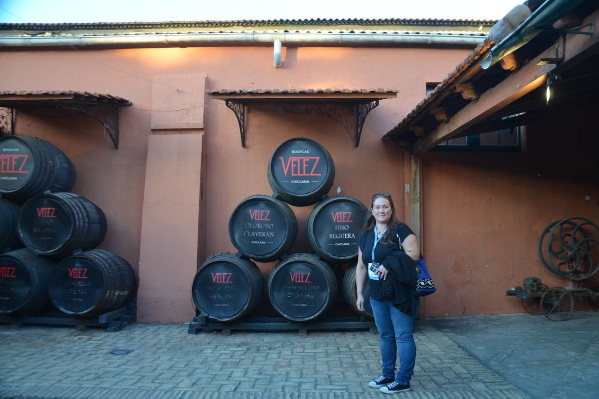Eine Frau steht vor einer Wand eines Gebäudes in Ockertönen, an der mehrere Weinfässer der Bodegas Velez aus Chiclana in einer Pyramidenform gestapelt sind.