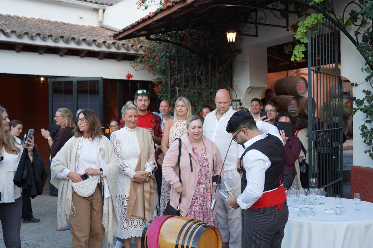 Eine Gruppe von Menschen beobachtet einen Kellner in traditioneller Kleidung, der Wein aus einem Fass in ein Glas einschenkt, in einem Innenhof eines Weinguts.