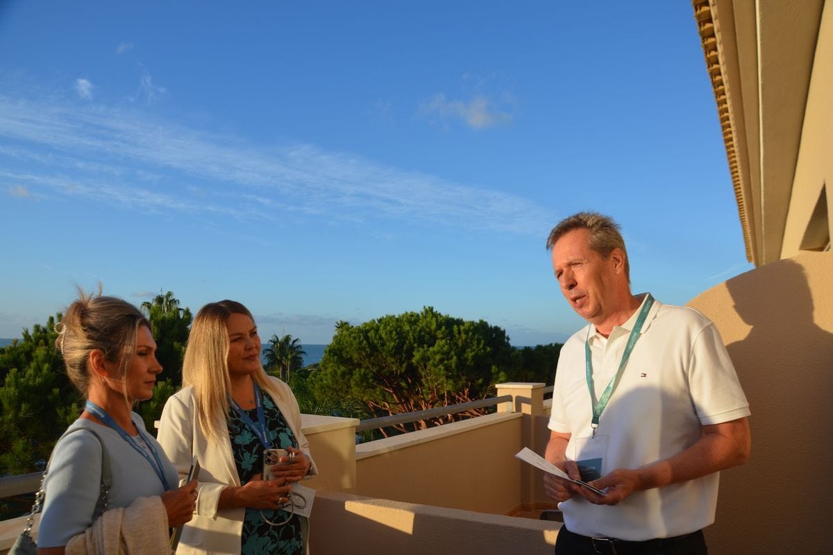 Drei Personen stehen auf einem Balkon vor einer Küstenlandschaft. Ein Mann spricht zu zwei Frauen, alle tragen Namensschilder an einem Umhängeband.